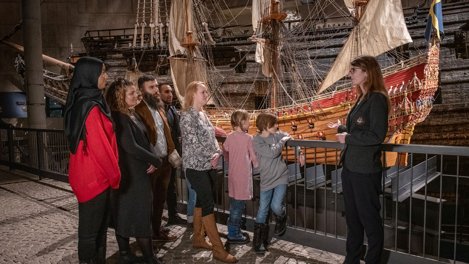 Blandad grupp av människor står framför en modell av skeppet Vasa, bakom dem syns även det riktiga skeppet i mörkt trä. Gruppen lyssnar på en kvinnlig guide med glasögon.m