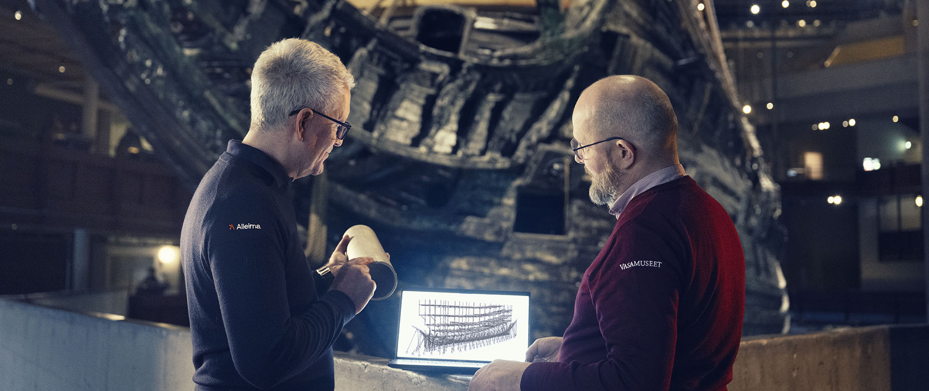 Två män framför skeppet Vasa i museet, de tittar på en datorskärm med ritning av skeppet i stöttning. 