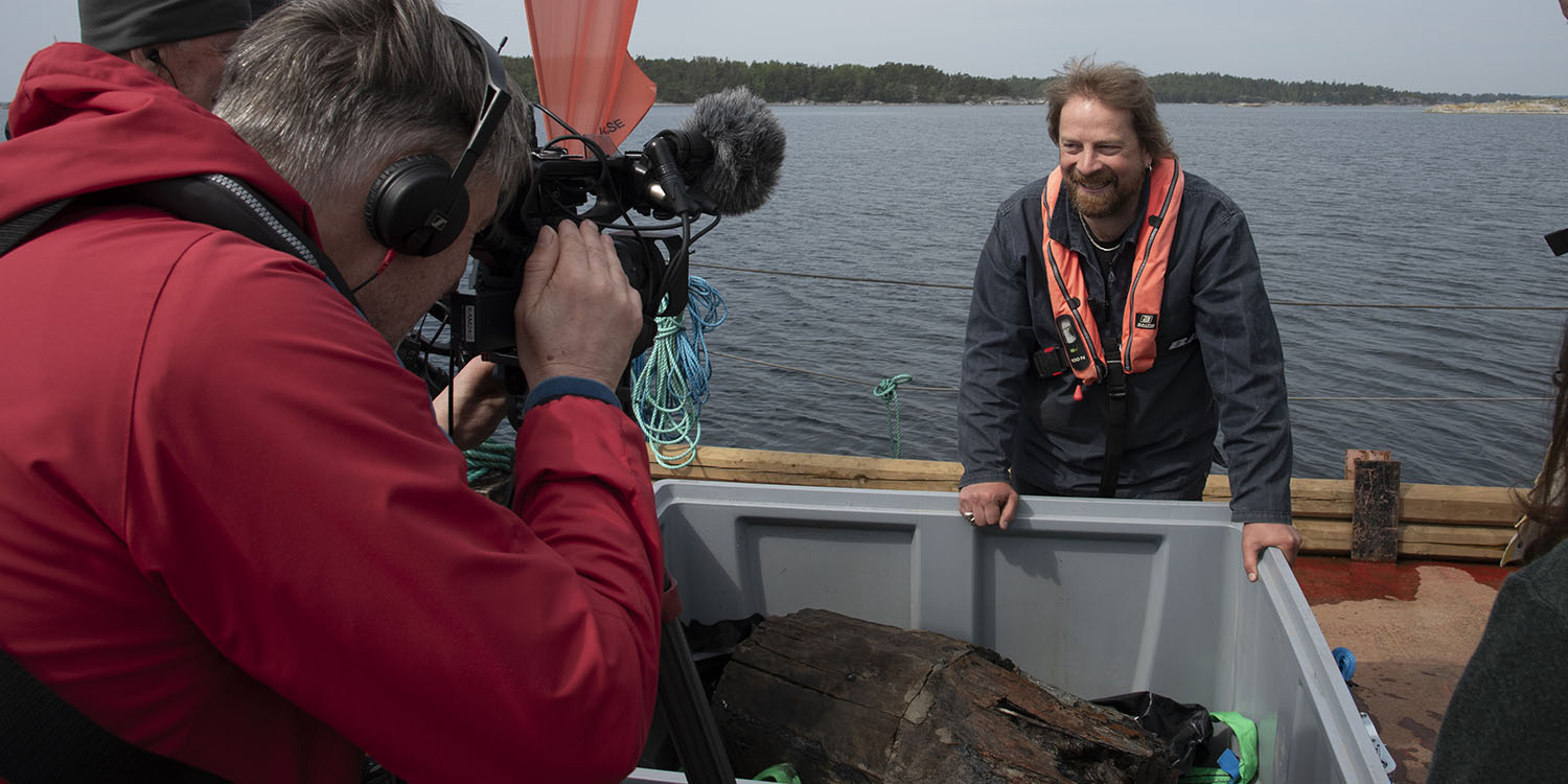 Pressuppbåd samlat runt tunna som ligger i grå låda, marinarkeolog JIm Hansson visar upp.